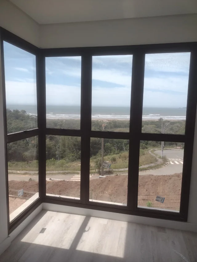 Ambiente interno com esquadria escura envidraçada de canto e piso com padrão de madeira.