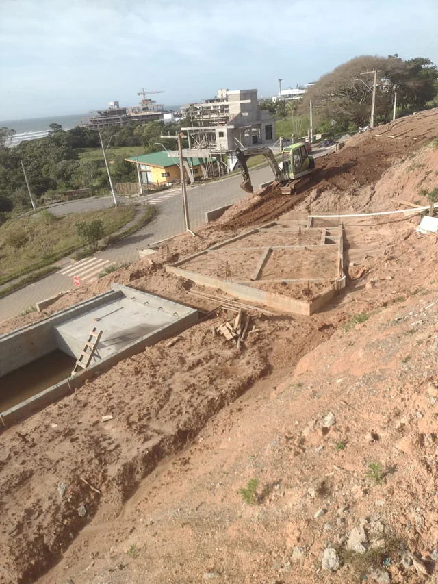 Terreno em declive com escavadeira, solo movimentado e estruturas baixas de concreto para fundação.