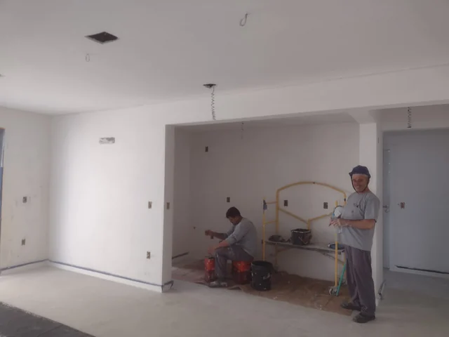 Dois trabalhadores em ambiente interno de paredes claras, com baldes, andaime e fiação aparente no teto.