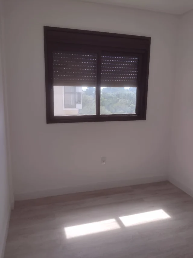 Cômodo de paredes claras com janela escura, persiana parcialmente fechada e piso com padrão de madeira.