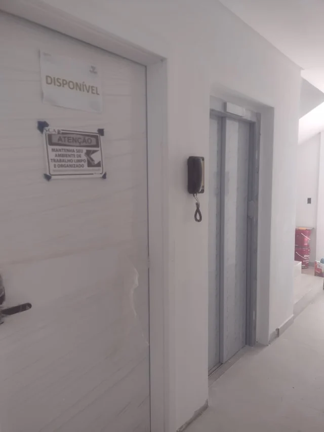 Corredor de paredes claras com porta protegida e sinalizada, interfone e outras portas ao fundo.