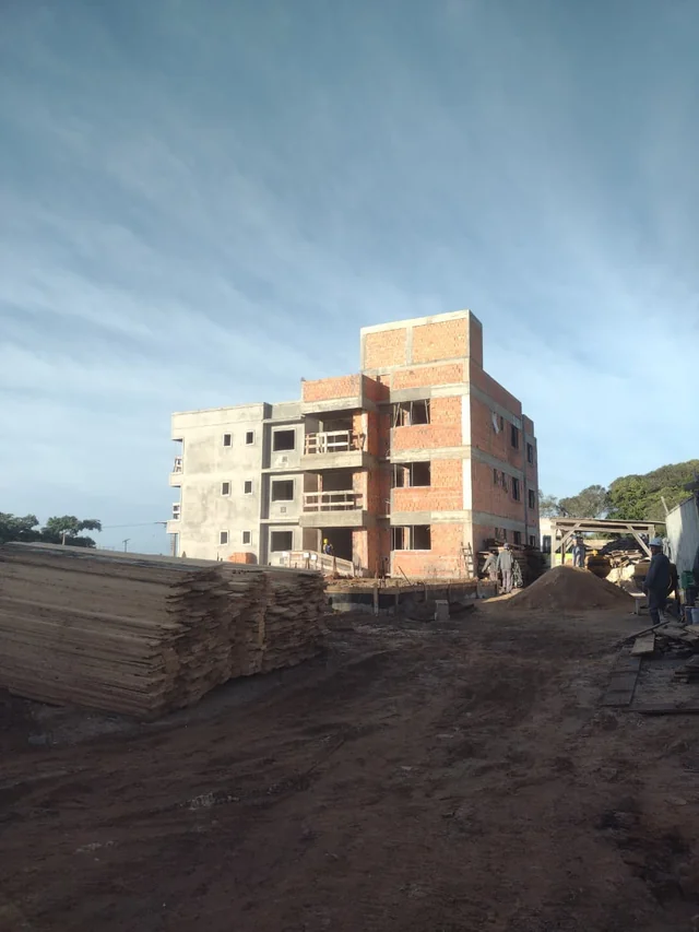 Edifício com estrutura de concreto, paredes de tijolos aparentes, vãos abertos e materiais no terreno.