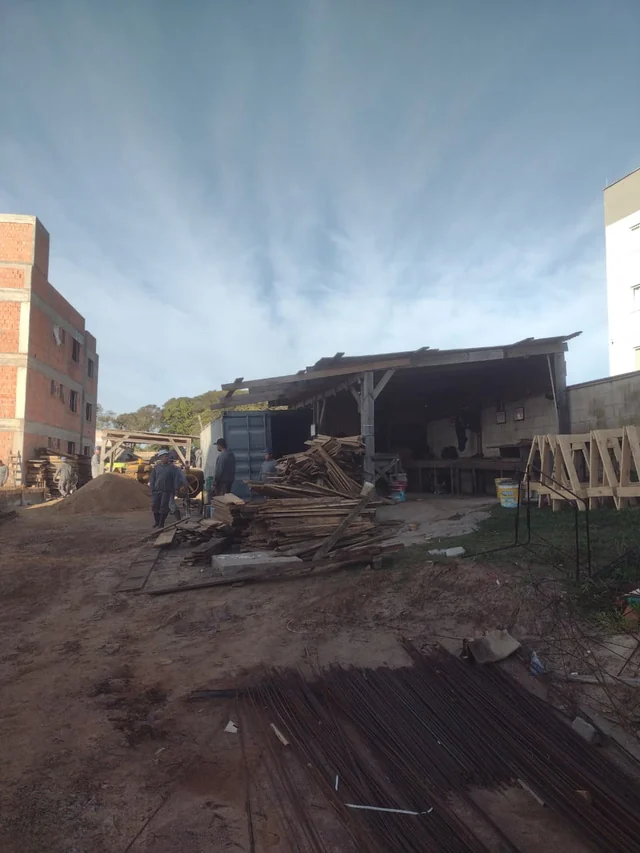 Canteiro com abrigo, pilhas de madeira, barras e armaduras de aço e trabalhadores.